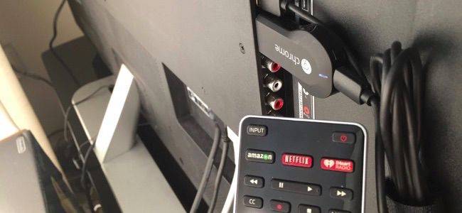 A remote control is in the foreground pointed at a Chromecast plugged into the back of a TV.