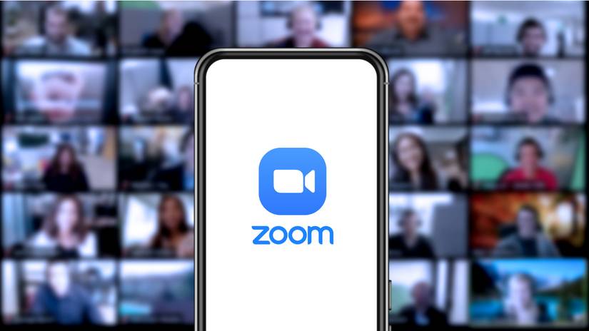 O logotipo do Zoom em um smartphone com uma videochamada em segundo plano.