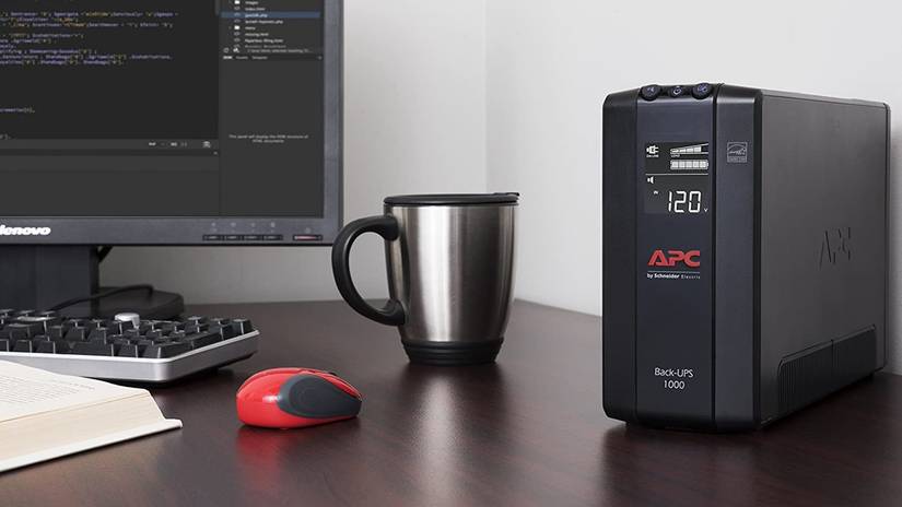 Um UPS APC sentado em uma mesa com monitor, mouse, teclado e caneca de café.