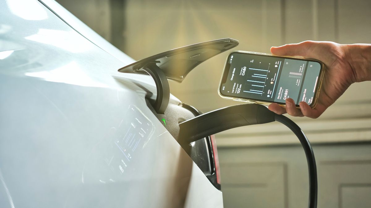 An electric car plugged into a charger station with a person holding a smartphone next to the connector.