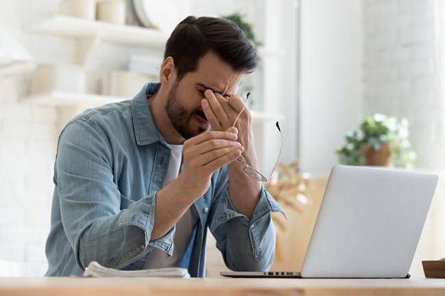 A man sitting in front of a laptop with eye strain, rubbing his eyes.