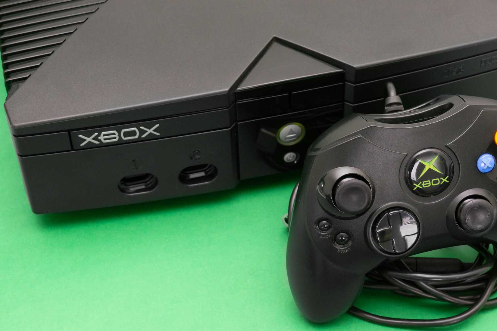 O console Xbox original da Microsoft ligado, em uma superfície verde e com um Controller S na frente.