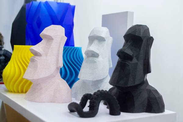 A collection of moai-inspired sculptures displayed on a white table. These stylized representations vary in color and form, including a pastel pink, a bright yellow, a light grey, and a deep black sculpture, all with the characteristic elongated noses and strong jawlines typical of moai statues. In the foreground is a small, intricate black sculpture resembling a curled tentacle or a looped object, providing a contrast in texture and shape to the smooth lines of the moai figures.