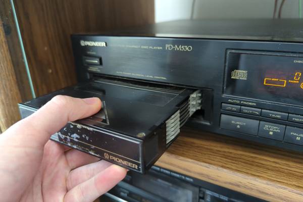 A hand pulls a multi-disc CD cartridge out of a CD player deck.