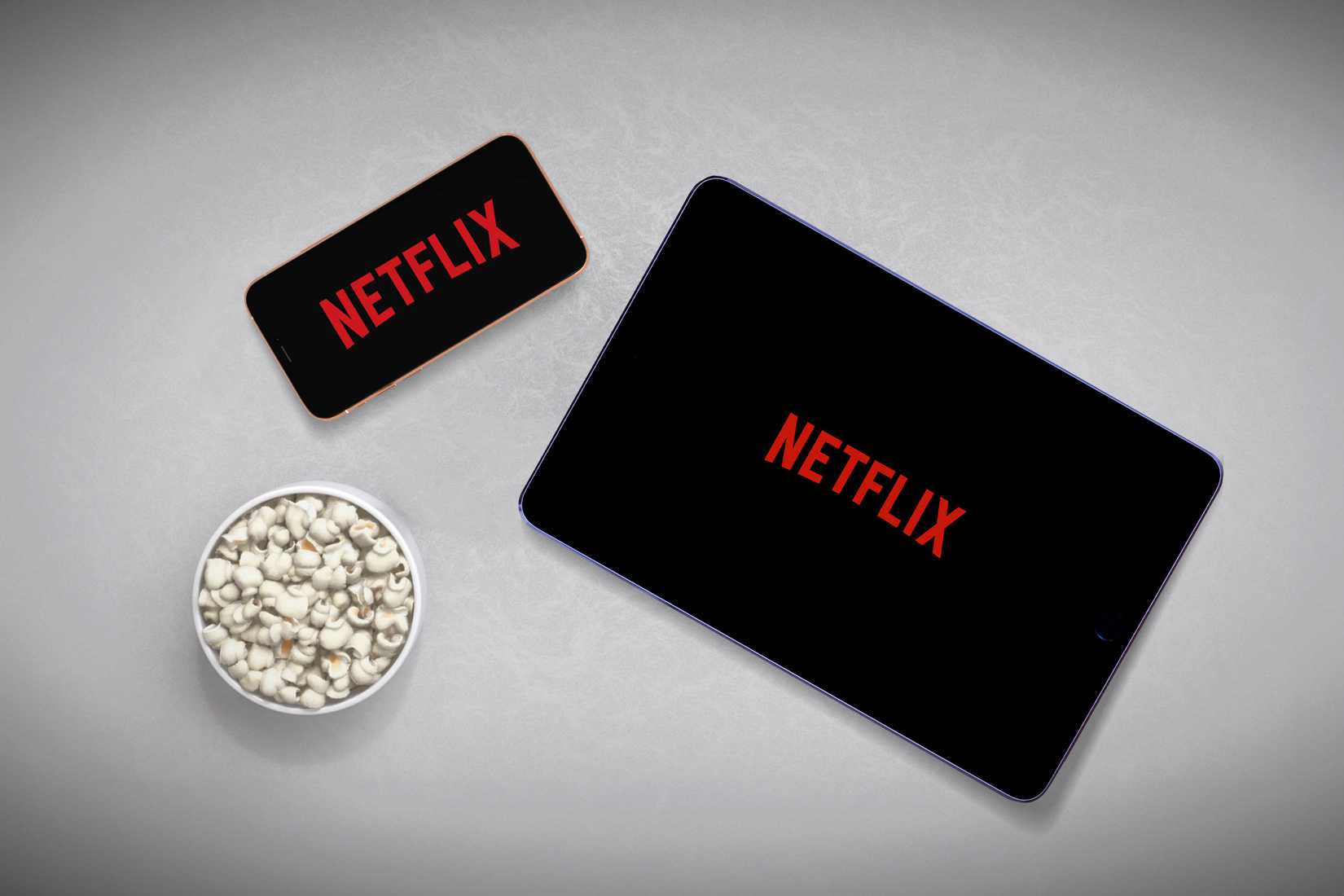 A smartphone on the left and a tablet on the right, both with the Netflix logo on the screen, and a bowl of popcorn in the bottom left corner.
