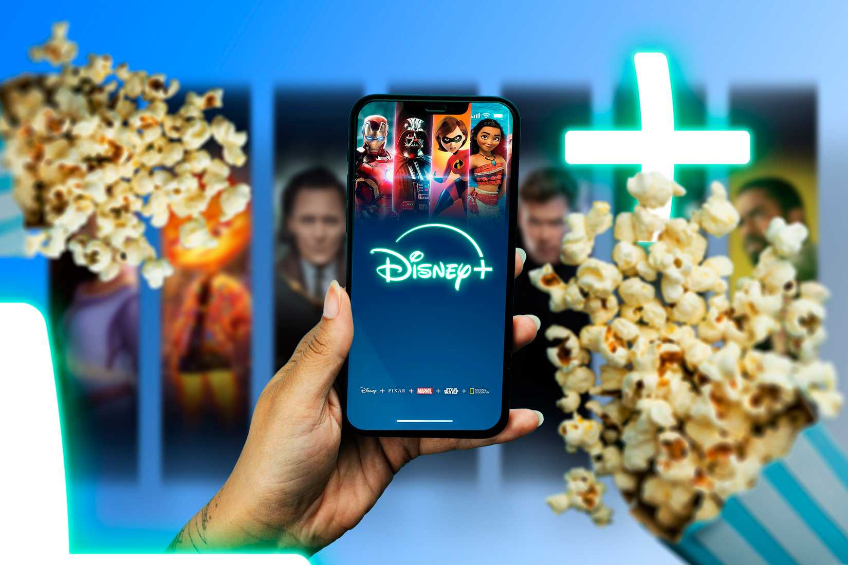 Рука держит смартфон с включённым экраном Disney+, рядом две пачки попкорна.