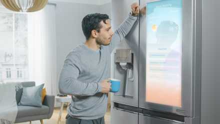 Man leaning against a smart fridge showing weather on the screen.