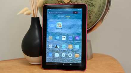 The Amazon Fire HD sitting on a wood-grain desk showing the device's home screen.