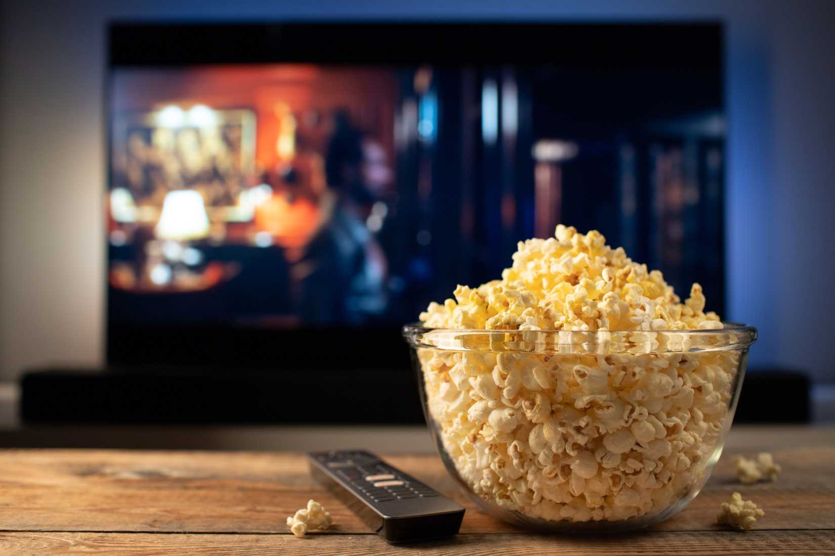 A glass bowl of popcorn and remote control while in the background the TV plays.