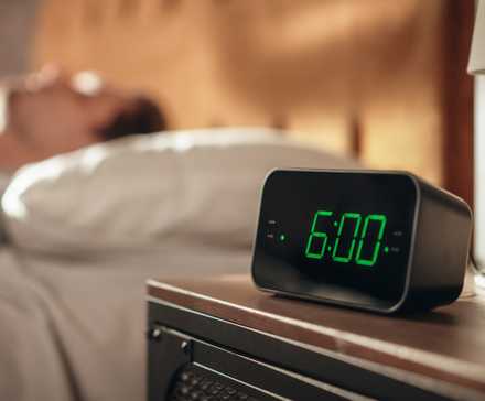 Мужчина в постели рядом с будильником, показывающим 6 утра.
