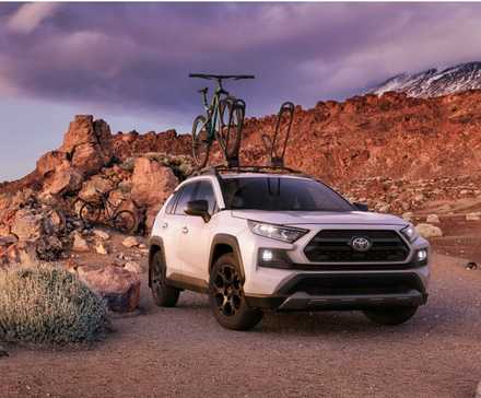 Белая Toyota RAV4 2020 года съезжает с асфальта с велосипедом на крыше.