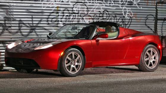 Full view of a red first-generation Tesla Roadster parked in front of metal roller shutters covered in graffiti. 