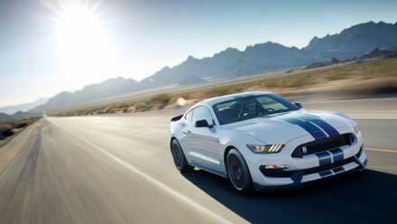 Front 3/4 view of a white 2017 Ford Mustang Shelby GT350 driving on a country road with mountains in the background.