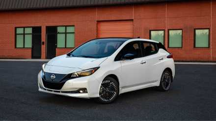 Front 3/4 view of a white 2025 Nissan Leaf parked on tarmac outside of a brick building.