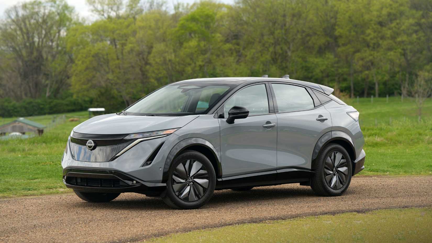 Front 3/4 view of a gray 2025 Nissan Ariya parked on a gravel track with grass and trees in the background.