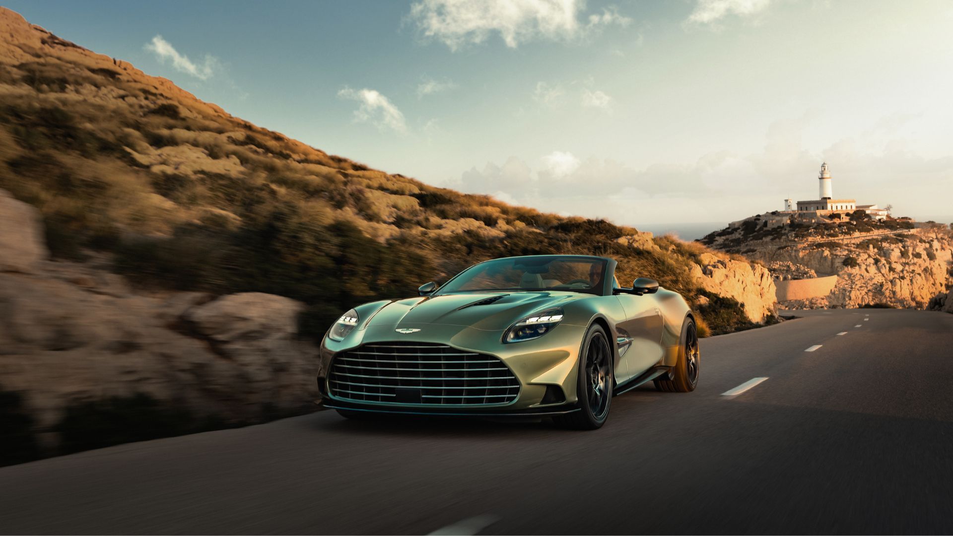 Front 3/4 view of the new Aston Martin Vanquish Volante driving on a coastal road with a lighthouse in the background.