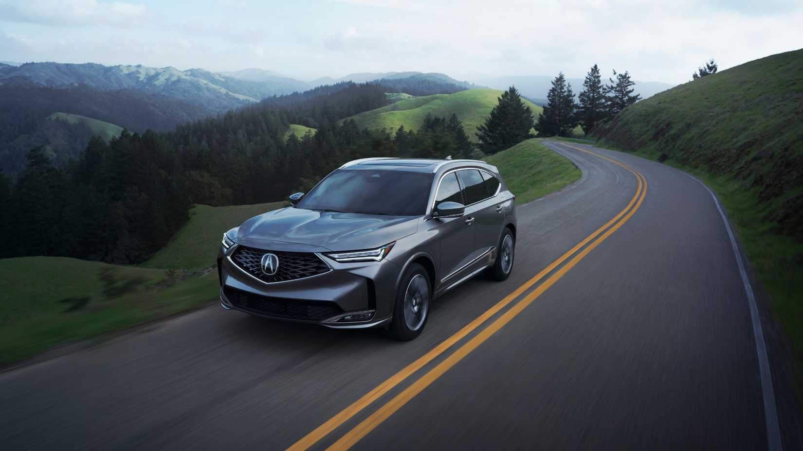 Front 3/4 view of a 2025 Acura MDX driving on a country road with mountains in the background.