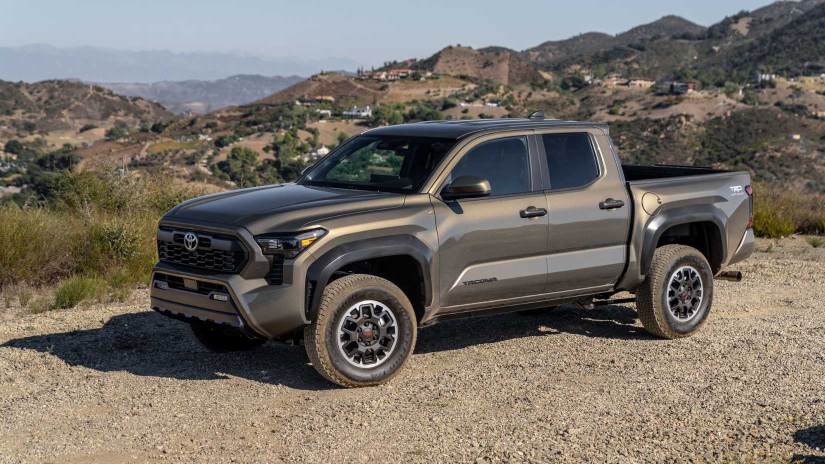 Front 3/4 view of a bronze 2025 Toyota Tacoma TRD parked on gravel with mountains in the background.