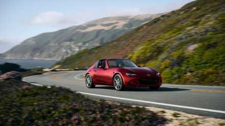 Front 3/4 shot of a red 2024 Mazda MX-5 Miata RF driving on a winding road with the ocean in the background.