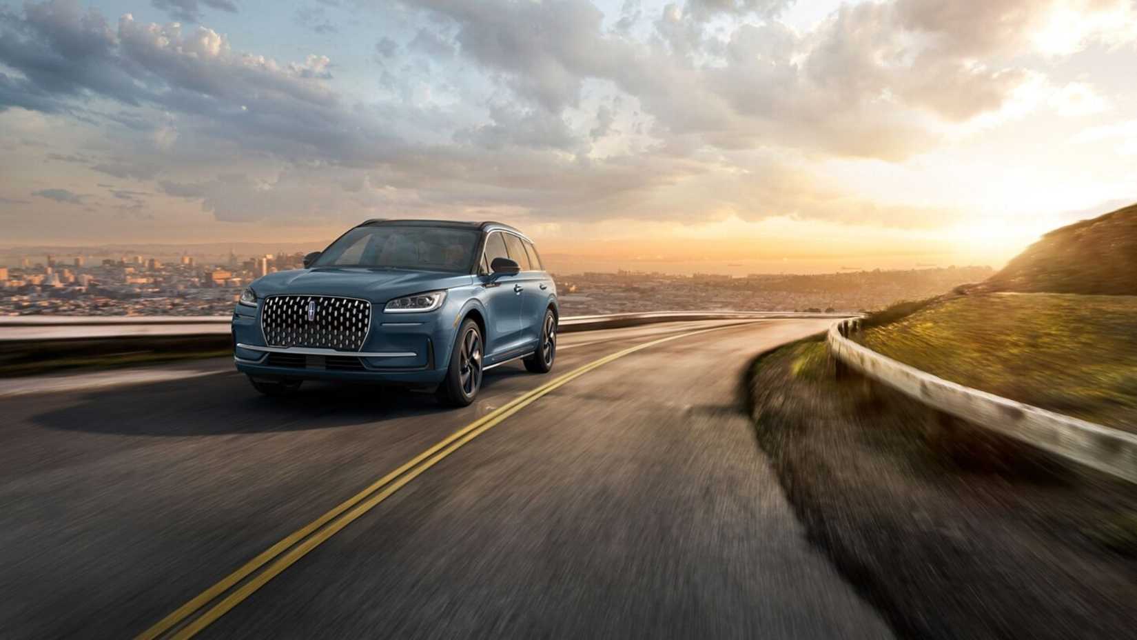 Dynamic front 3/4 shot of a blue 2025 Lincoln Corsair Grand Touring driving on a winding road with a city landscape in the background.