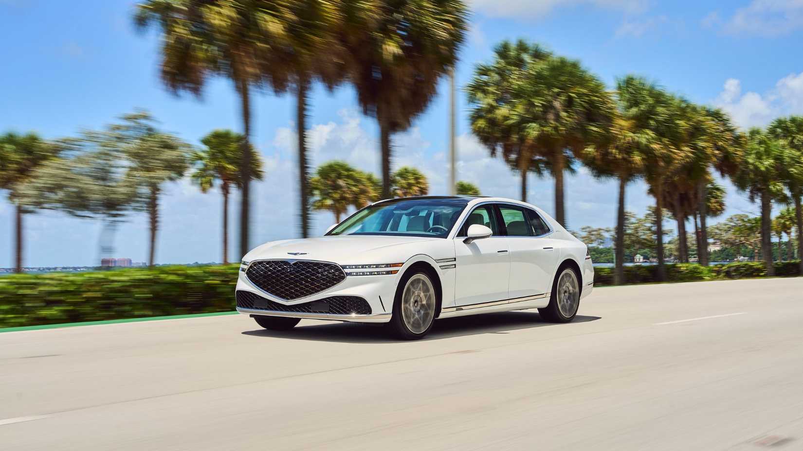 Dynamic front 3/4 shot of a white 2024 Genesis G90 driving on a highway with palm trees in the background.