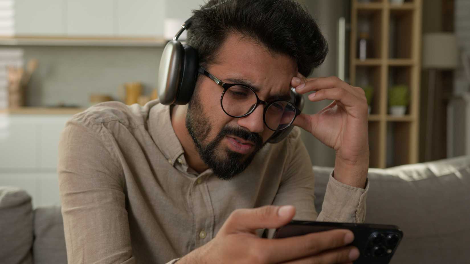 A frustrated man looking at an iPhone with AirPods Max headphones on.