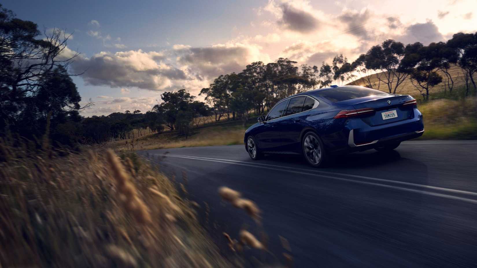 Rear 3/4 view of a blue 2024 BMW 5 Series driving on a country road with trees in the background.