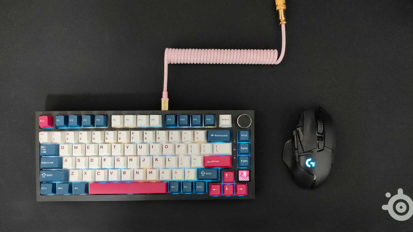 A mechanical keyboard with a coiled pink cable and a Logitech G502 wireless mouse on a black SteelSeries QcK Performance mousepad.