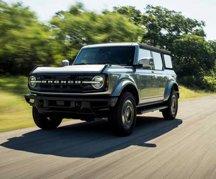 Dynamic front 3/4 shot of a 2021 Ford Bronco driving on a country road lined by trees.