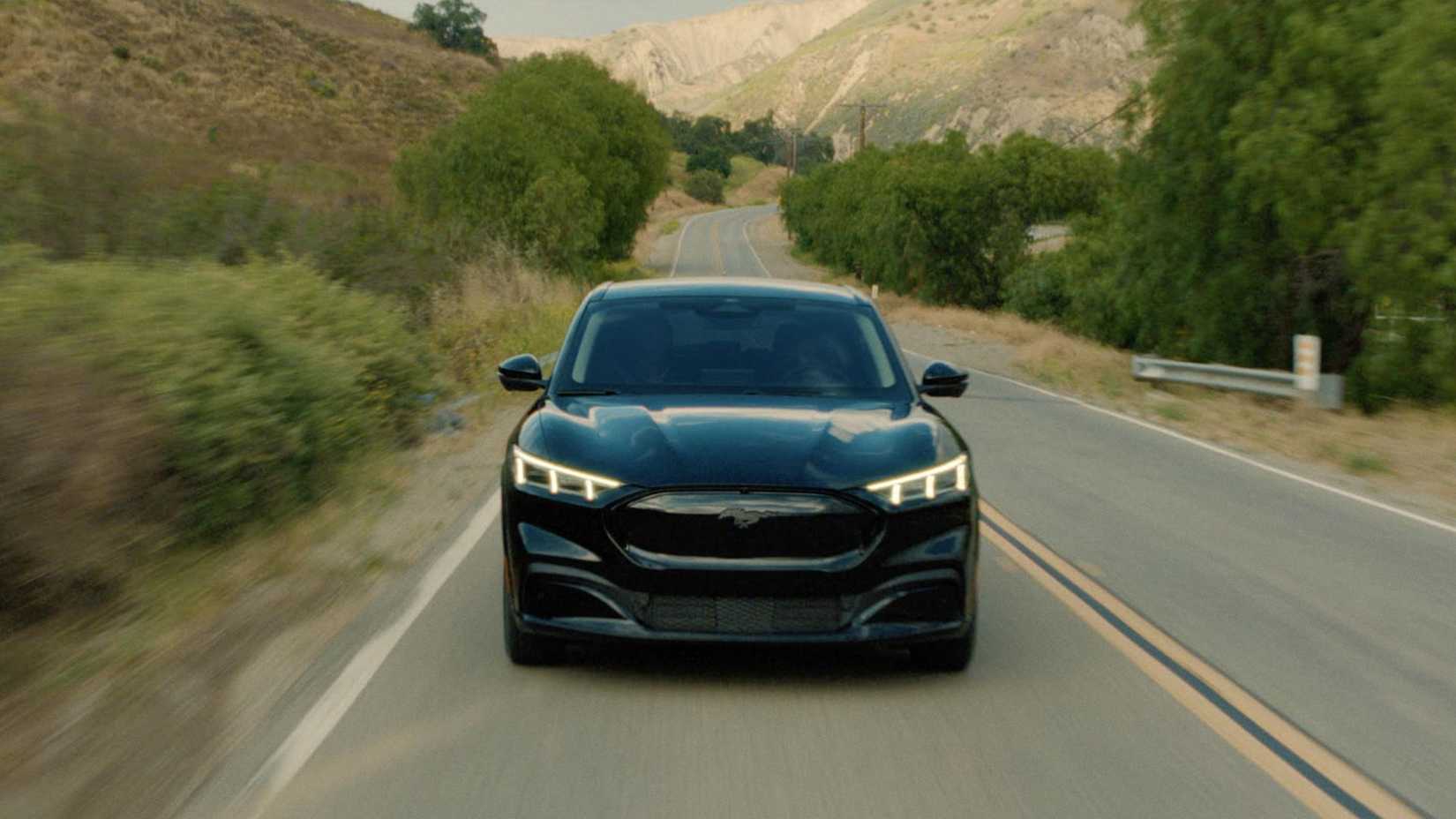 Toma dinámica de la parte delantera de un Mustang Mach-E Premium 2023 negro conduciendo por una carretera rural.