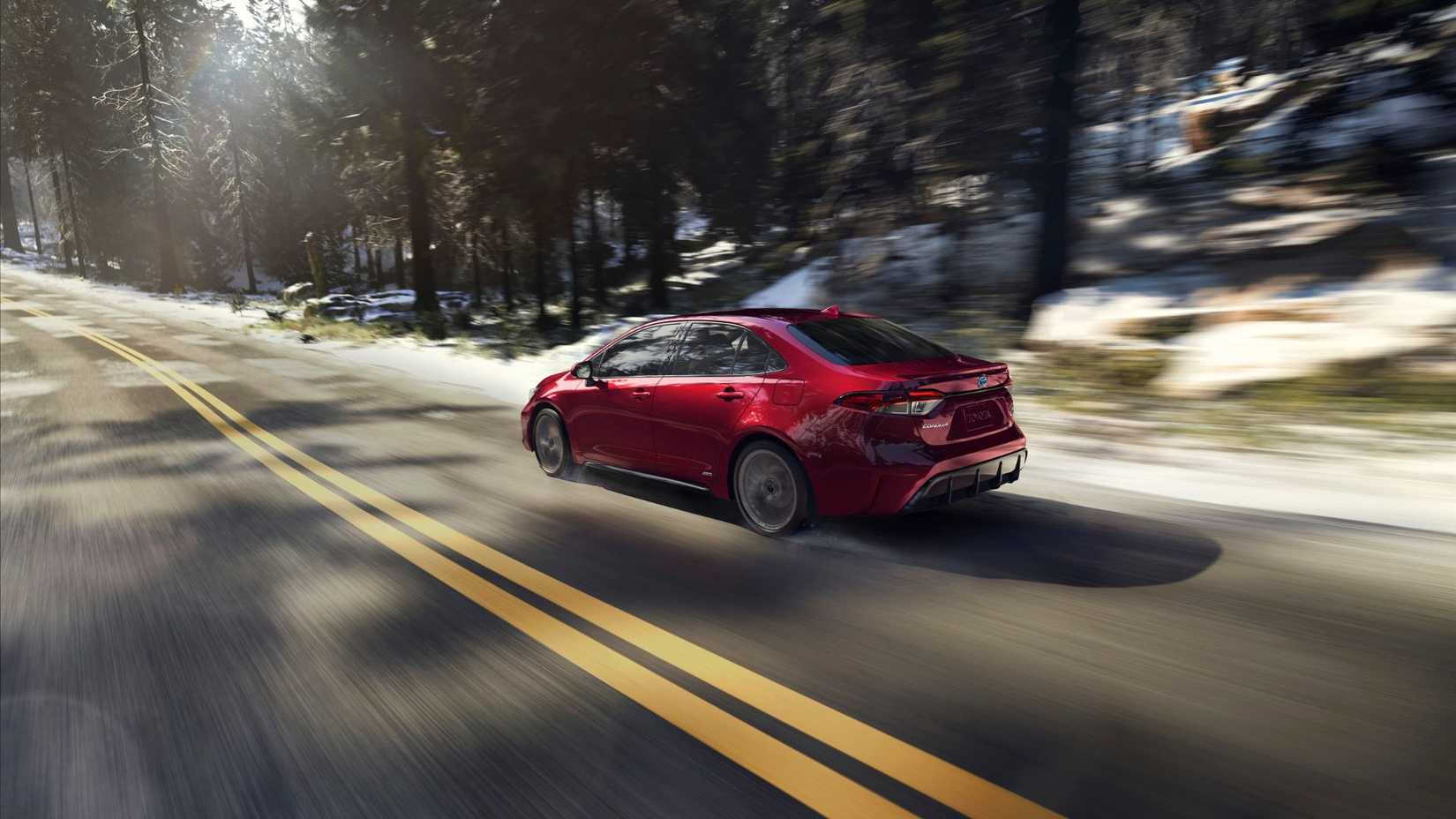 Dynamic rear 3/4 shot of a red 2025 Toyota Corolla Hybrid driving on a country road with trees in the way.
