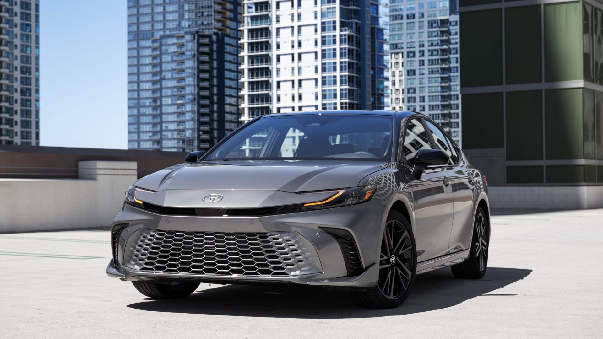 Static front 3/4 shot of a gray 2025 Toyota Camry XSE AWD parked with highrise buildings in the background.