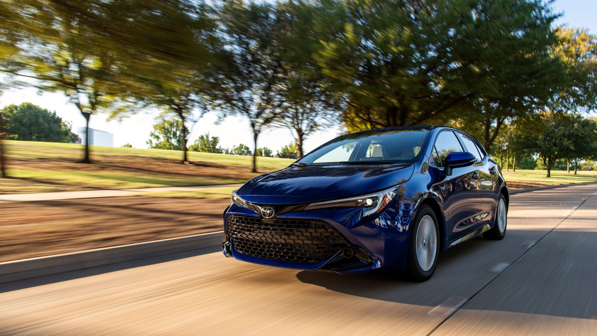 Dynamic front 3/4 shot of a blue 2025 Toyota Corolla Hatchback SE driving on a tree-lined road.
