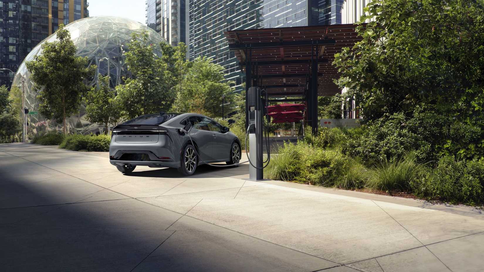 TOMA ESTÁTICA TROPA 3/4 de un Toyota Prius PHEV gris 2025 conectado en un punto de carga fuera de un edificio moderno en una ciudad.