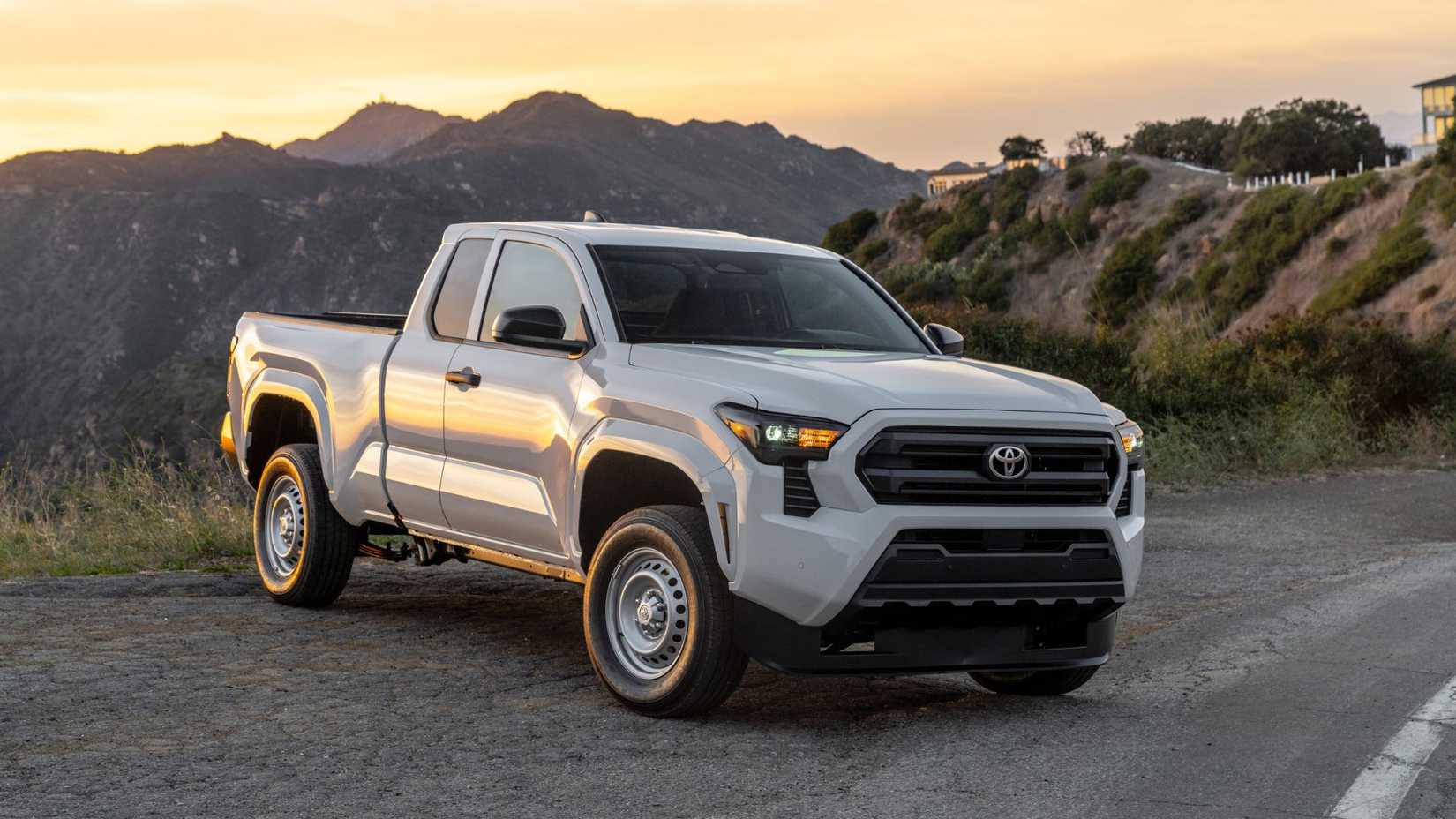 Static 3/4 front shot of a white 2025 Toyota Tacoma SR with mountains and a property in the background.