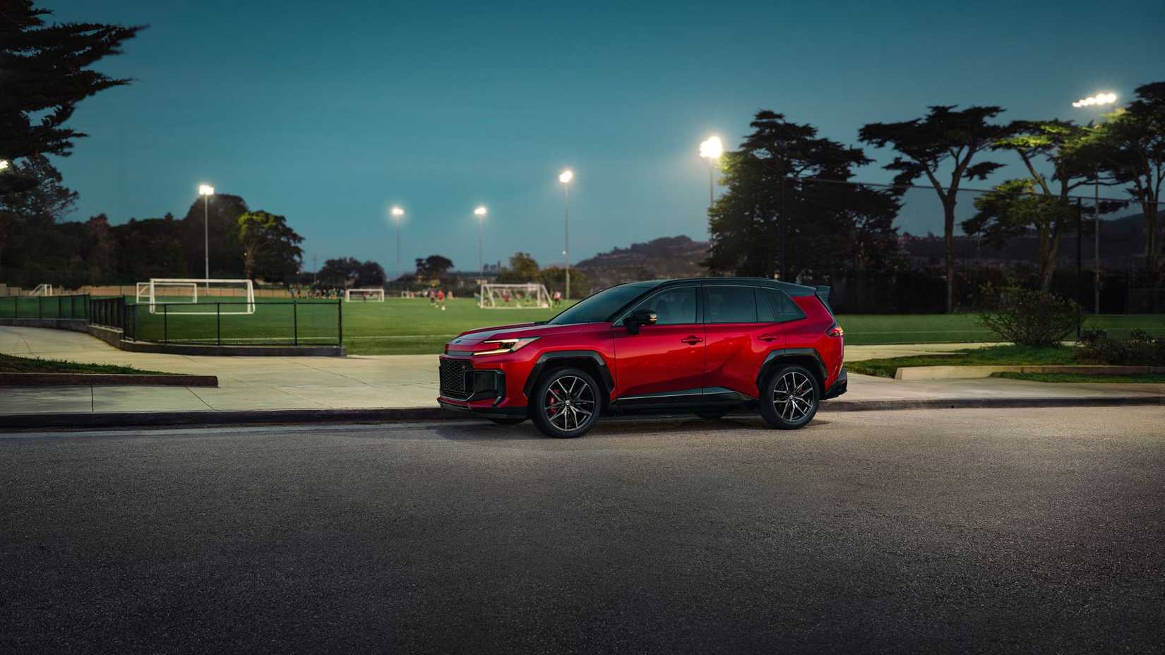 Static side profile shot of a red 2026 Toyota RAV4 PHEV GR Sport parked next to a soccer field.