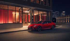 Static front 3/4 shot of a red 2026 Toyota Camry Nightshade parked outside of a building.