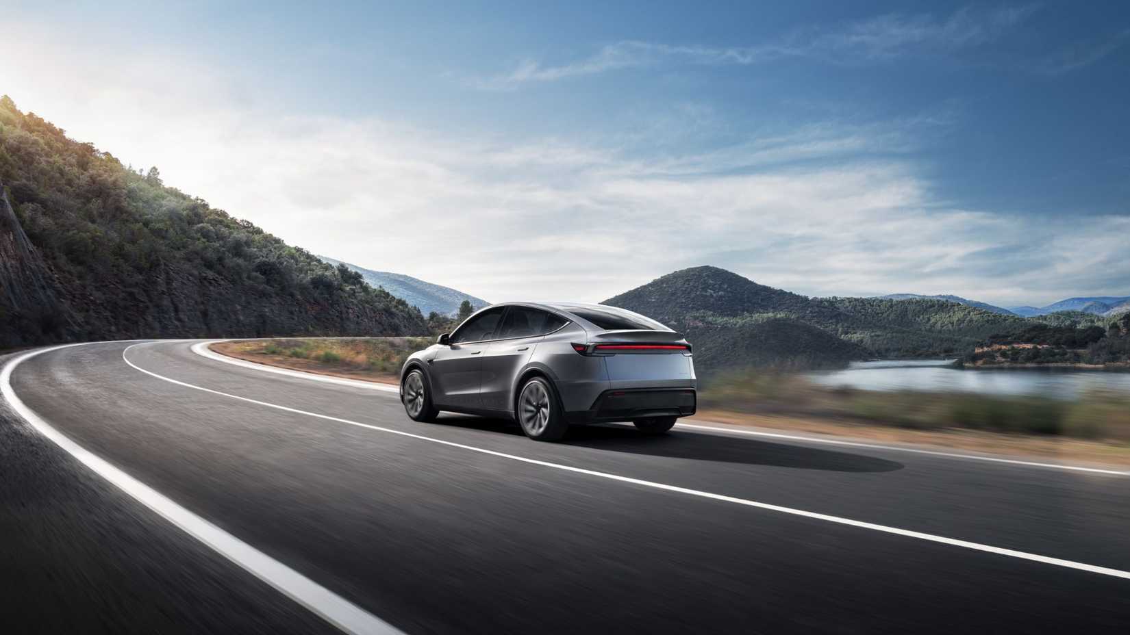 Tesla Model Y движется по загородной дороге вдоль озера, вид сзади.