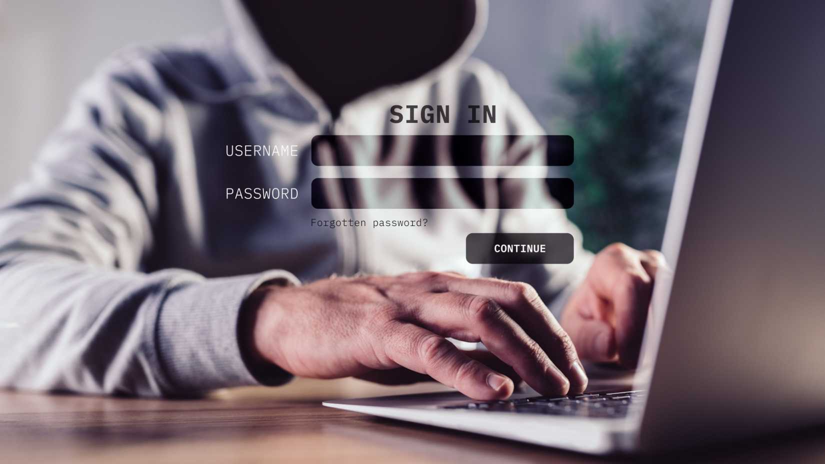 A dark, hooded figure is ominously using a laptop.