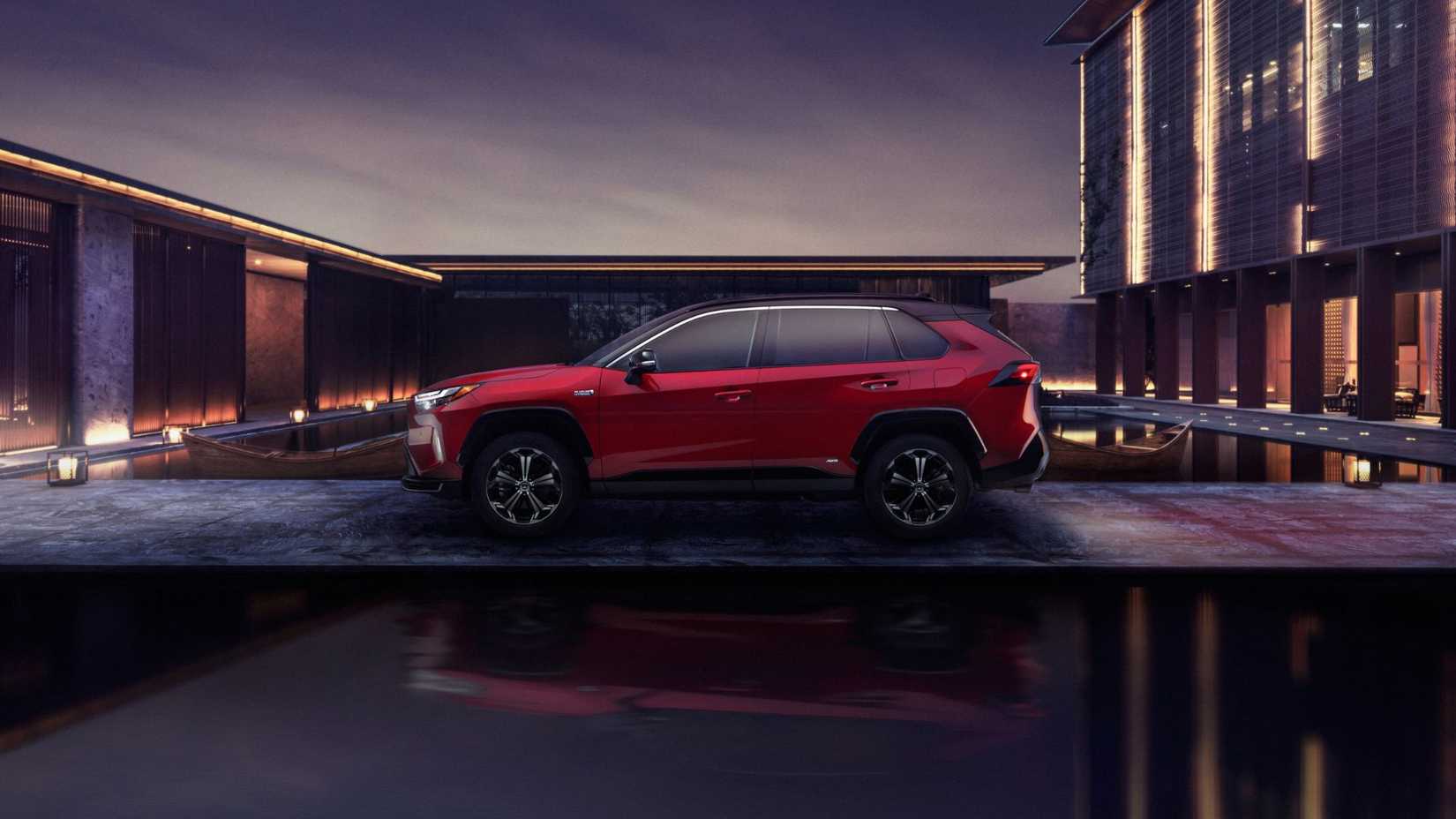 Static side profile shot of a red 2024 Toyota RAV4 Prime XSE at night with modern buildings in the background.