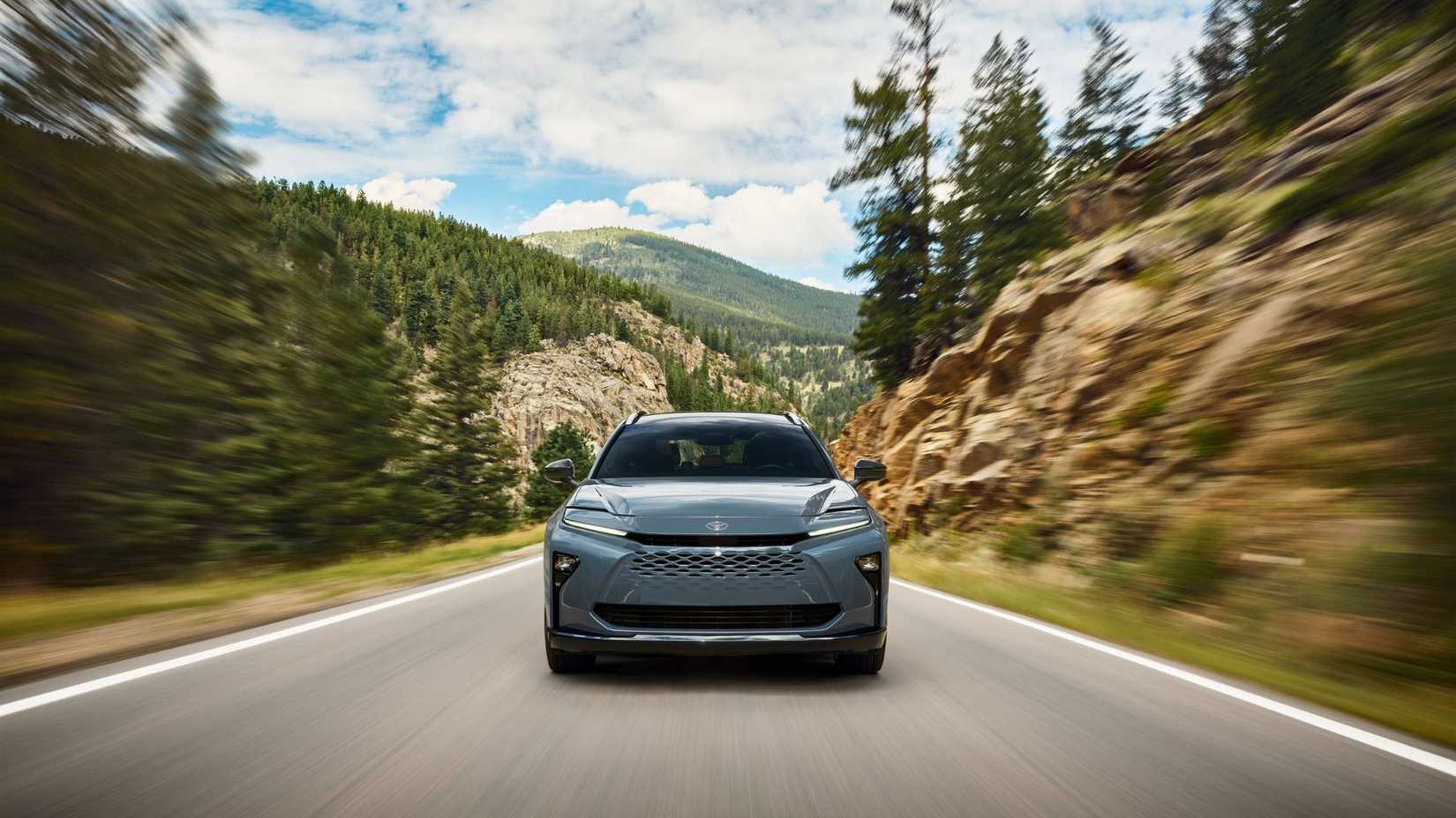 Dynamic front-end shot of a gray 2025 Toyota Crown Signia driving on a country road lined by trees a mountain in the background.