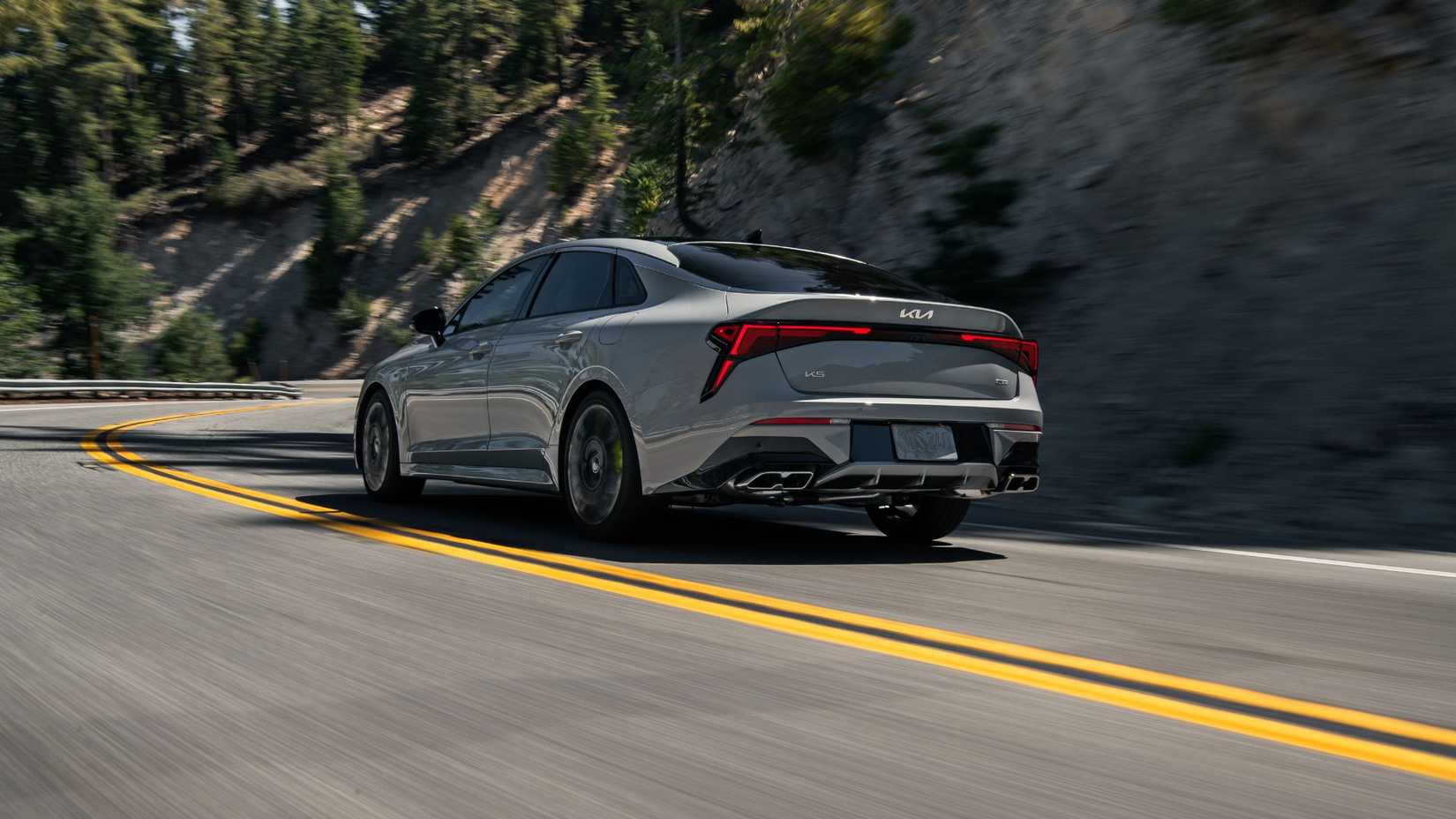 Dynamic rear 3/4 shot of a gray 2025 Kia K5 driving on a country road through the mountains.