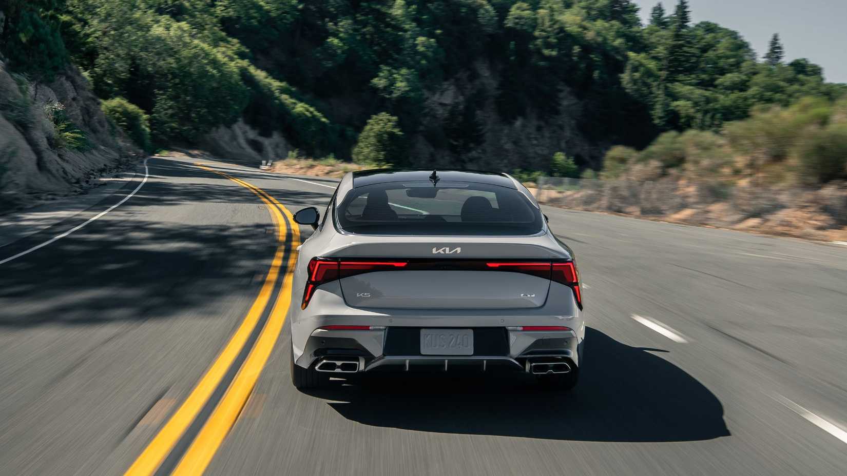 Toma dinámica de la parte trasera de un Kia K5 gris 2025 conduciendo por un camino rural a través de las montañas.