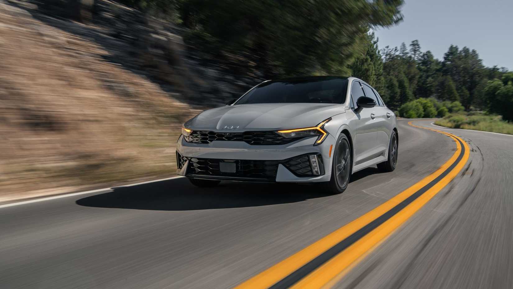 Dynamic front 3/4 shot of a gray 2025 Kia K5 driving on a country road through the mountains.