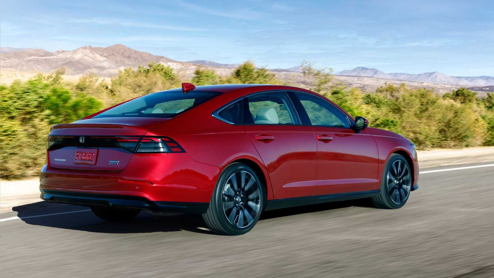 Dynamic rear 3/4 shot of a red 2023 Honda Accord Touring driving on a country road with mountains in the background.