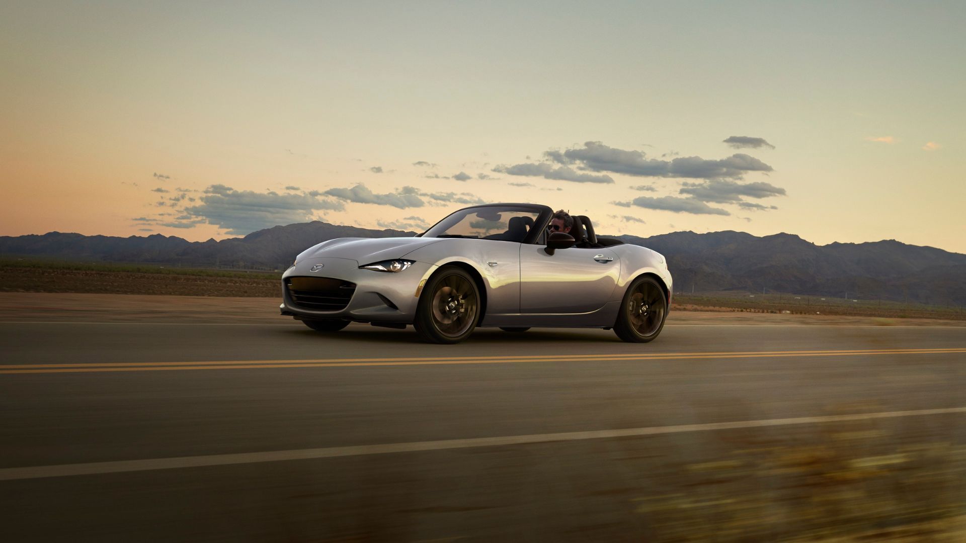 Dynamic front 3/4 shot of a silver 2024 Mazda MX-5 driving on a counattempt road with mountains in the background.