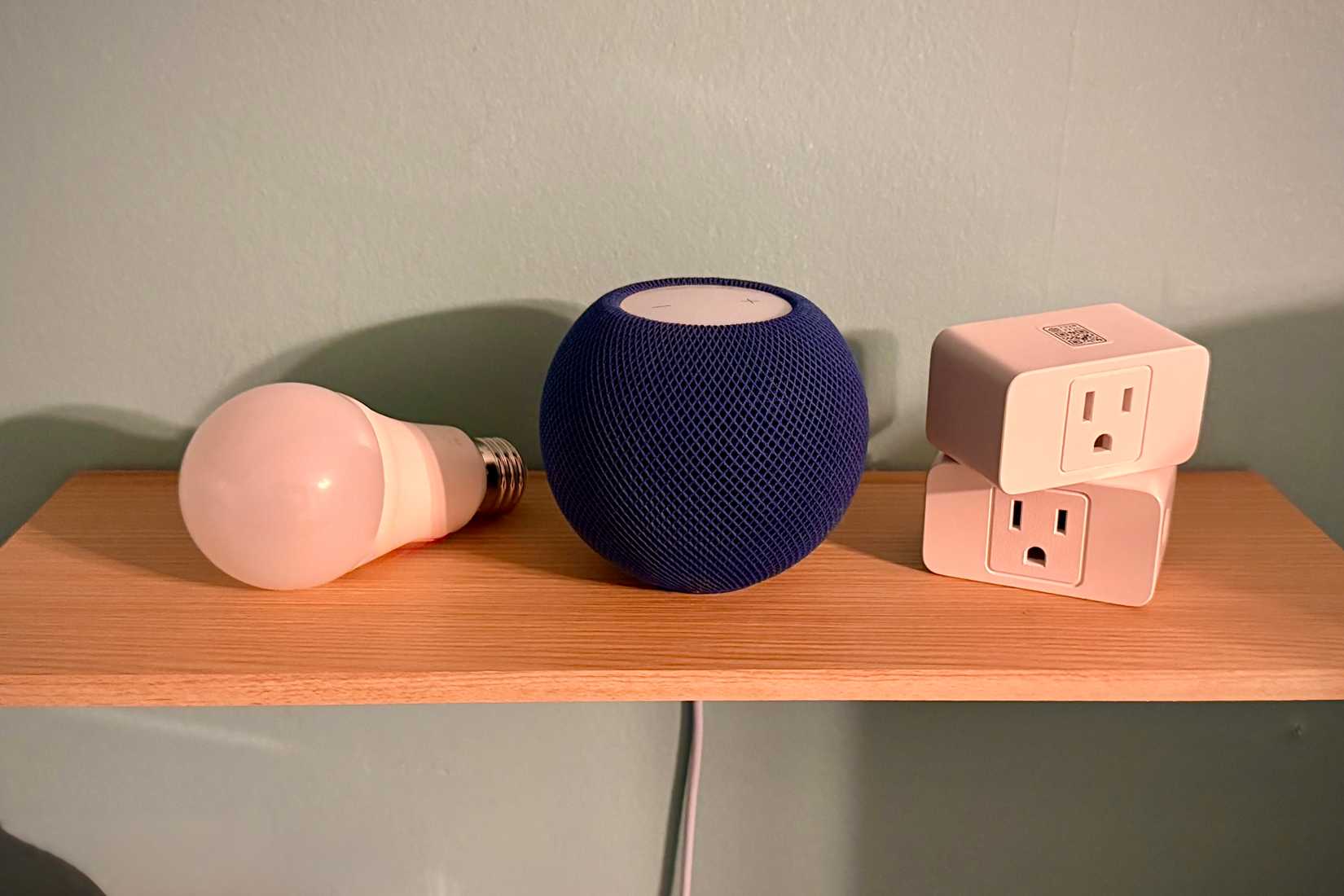 Bleu HomePod mini next to two smart plugs and a smart lightbulb on a shelf.