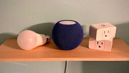 Bleu HomePod mini next to two smart plugs and a smart lightbulb on a shelf.