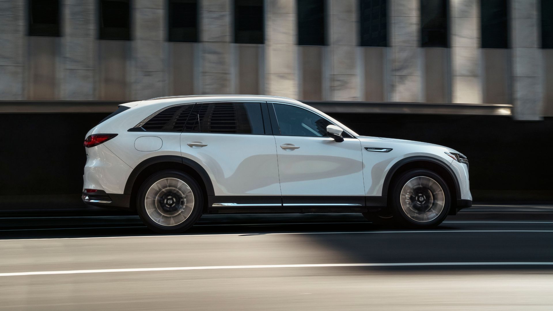 Dynamic side profile shot of a white 2025 mazda CX90 ‍driving in a city.