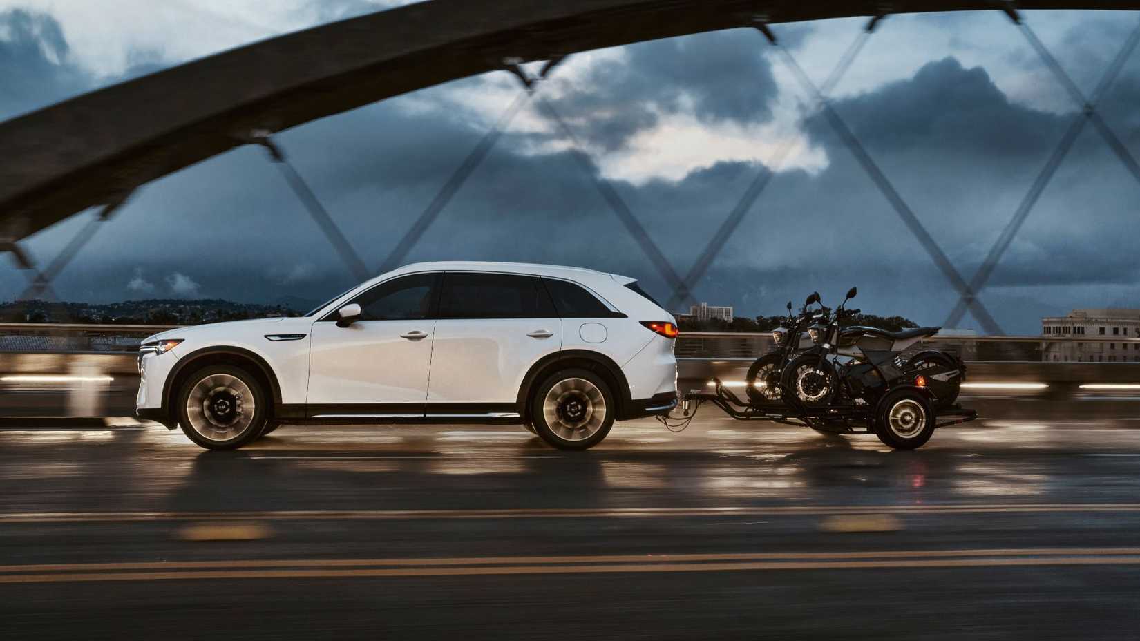 Dynamic side profile shot of a white 2025 Mazda CX-90 towing a trailer with motorbikes on over a bridge in a city.